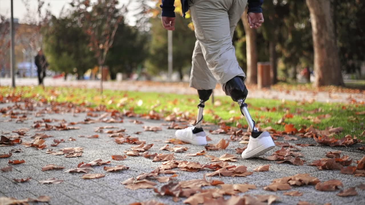 Slow motion view of a walking man with prosthetic legs and white sneakers in a park. Fallen yellow leaves on the ground