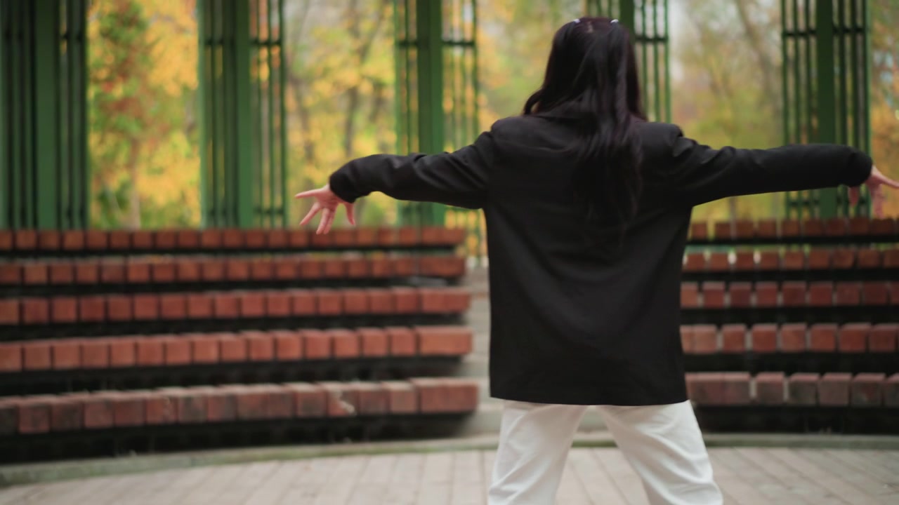 Young woman performs dance routine in outdoor park setting moving gracefully with her arms and legs. Woman wears black top and white pants against backdrop of red brick steps and green