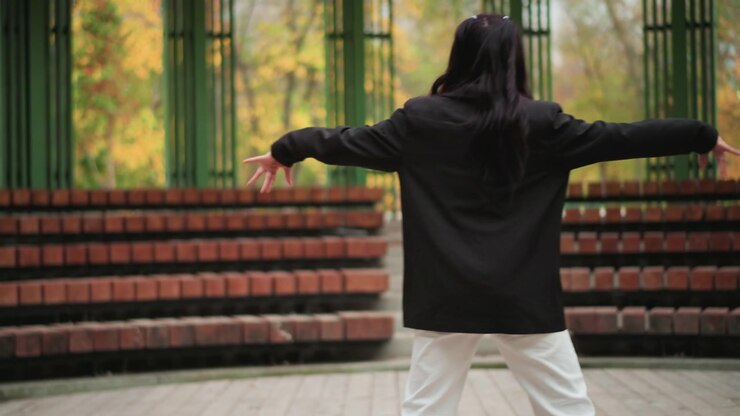 Young woman performs dance routine in outdoor park setting moving gracefully with her arms and legs. Woman wears black top and white pants against backdrop of red brick steps and green