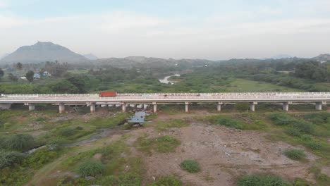 vista aérea sobre un vasto campo verde en la india, volando por encima de un puente de carretera con camiones conduciendo en él, tiro ancho, concepto de transporte