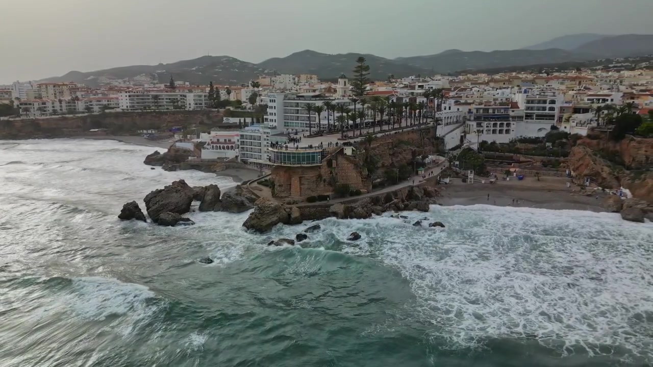 Luftfoto med drone af kystbyen Nerja med udsigtspunktet Balcón de Europa, klippekyster og voldsomme brændinger, hvide bygninger og grønne bakker i baggrunden, overskyet dagslys.