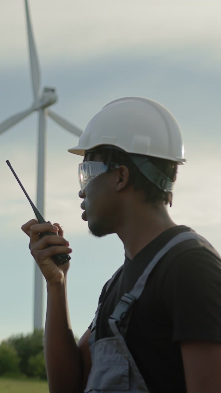 Man in hard hat and overalls uses radio near wind turbines monitoring operations in renewable energy