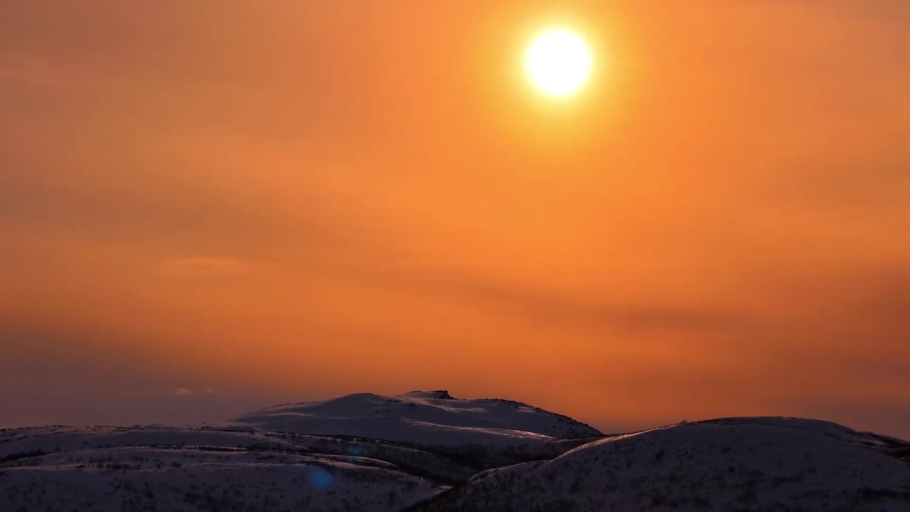 Gorgeous aerial drone footage from Giemaš mountains in Norway where a large orange sun is rising or setting with a vibrant colored orange sky lighting up the snow covered mountains below.