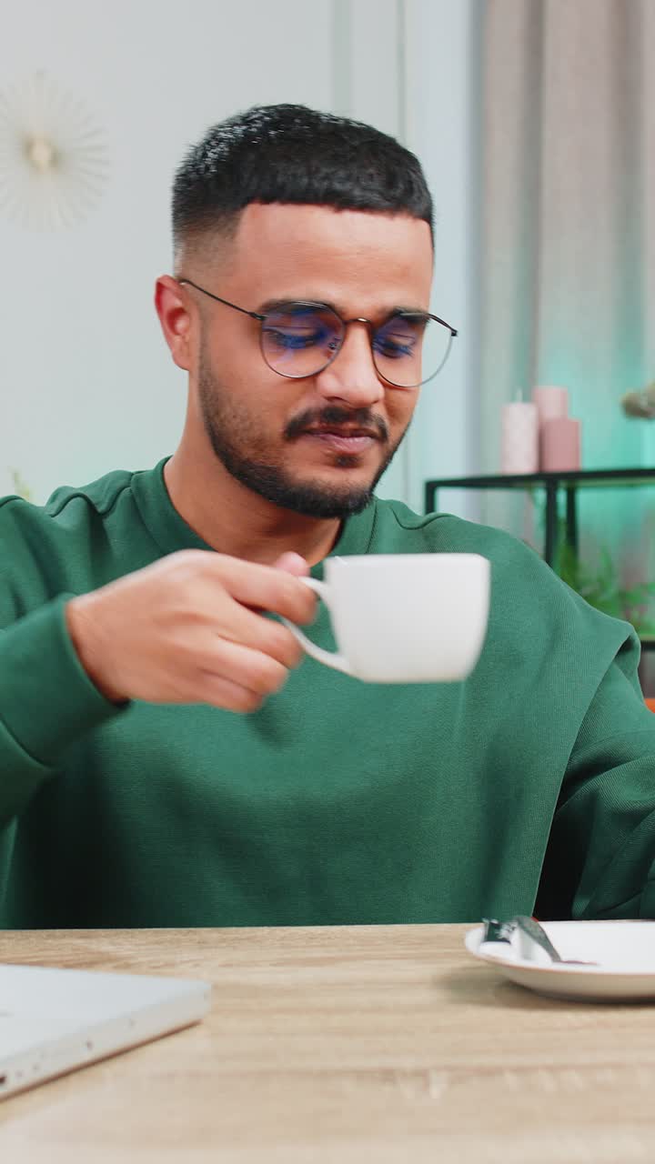 Mand, der arbejder på bærbar computer og drikker en kop varm kaffe, sidder ved bordet på en arbejdsplads i hjemmekontoret