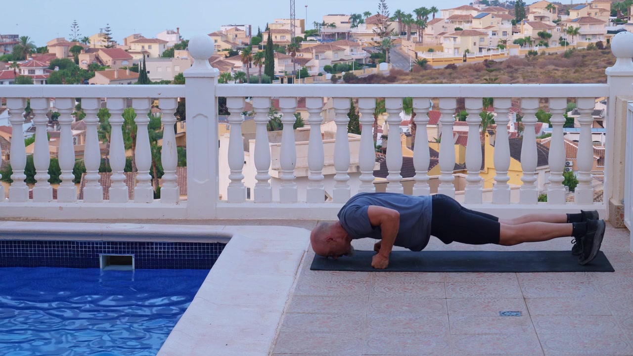 Side profile shot of a mature athletic man performing intense burpees and knuckle push ups on a fitness mat on a villa terrace next to a pool.
