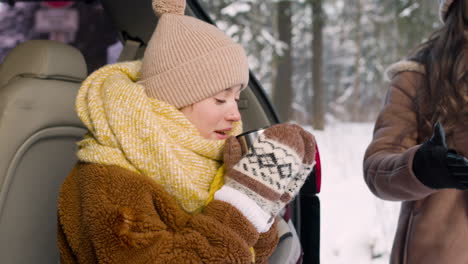 teenage girl dressed in winter clothes sitting in the trunk of a car drinking hot drink 1