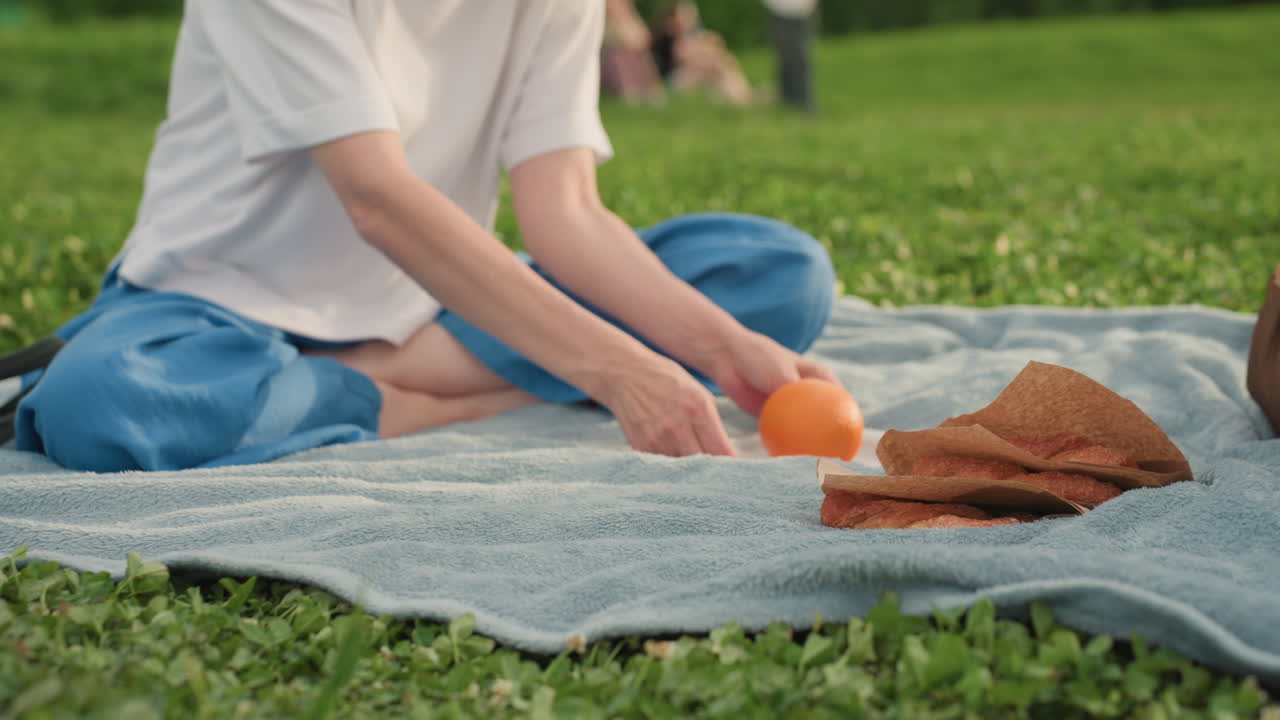 woman on picnic mat placing bowl with orange into bag while sitting on grass, sandwich and snacks nearby, summer park relaxation moment with casual outdoor preparation