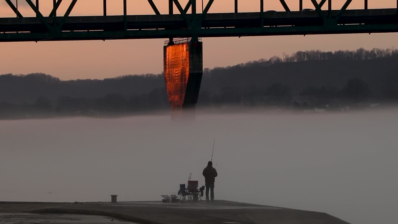 Lone fisherman is dwarfed by massive bridge while fishing in fog-covered river