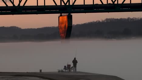 lone fisherman is dwarfed by massive bridge while fishing in fog-covered river
