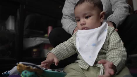 cute 4 month old baby wearing dribble bib playing with toy on the floor whilst being held so he doesn't fall back