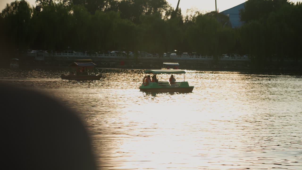 Scena lacustre al tramonto con una piccola barca che scivola sull'acqua scintillante, figure in silhouette che si godono una tranquilla remata contro la riva alberata e lo skyline distante, calda luce dorata e leggere increspature che creano.