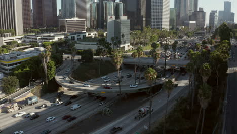 Big-Freeway-Highway-intersection-with-car-traffic-and-palm-trees-next-to-Los-Angeles-Downtown-Skyscraper-Skyline-with-beautiful-blue-sky,-Aerial-dolly-in-tilt-down-establisher