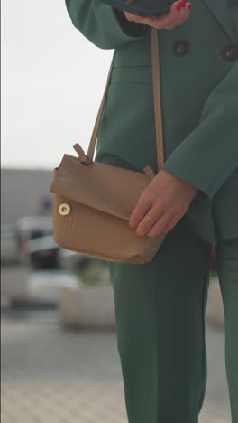 young woman in business suit receives call on phone and takes out gadget from cross-body handbag. female talks on phone walking about city on blurred background closeup