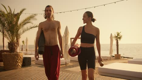 a brunette guy in red pants and a brunette girl in a black sports summer uniform are walking along a beach covered with boards and talking after a yoga class