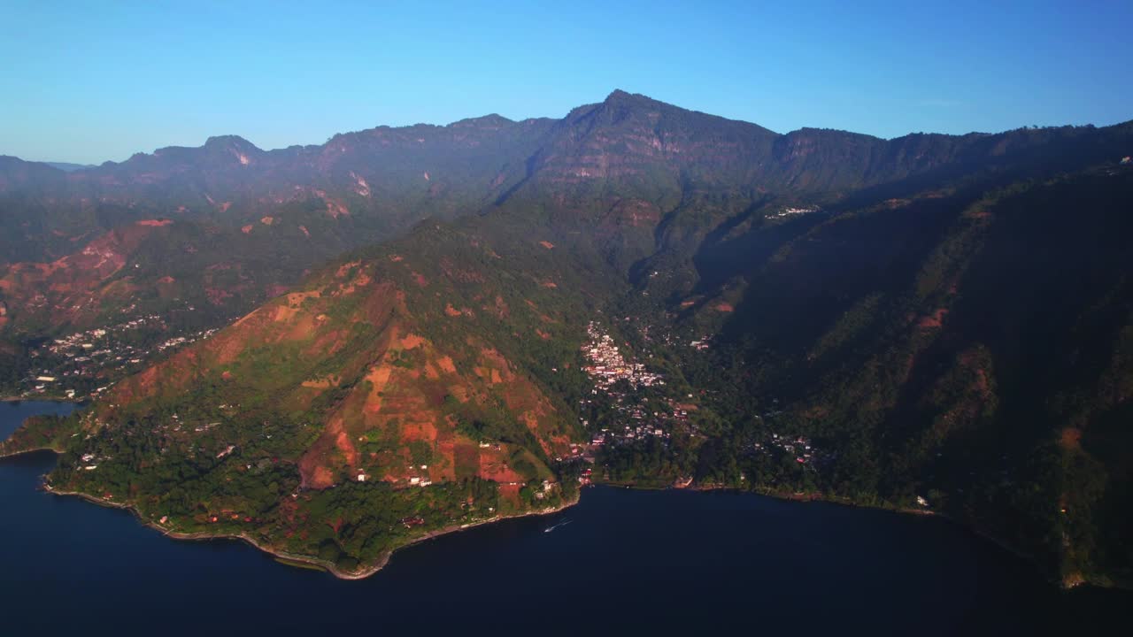 Drone che zooma allontanandosi, rivelando un villaggio in riva al lago, ripidi pendii vulcanici e acque blu intenso che formano una drammatica composizione naturale