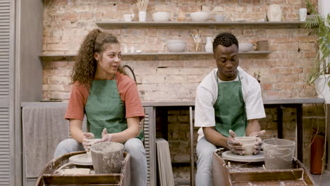 vista frontal de los empleados que usan delantal verde modelando piezas de cerámica en una rueda de alfarero en un taller mientras hablan entre ellos