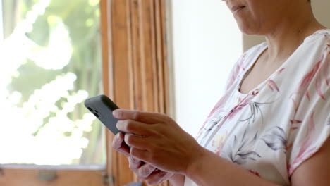 Nachdenkliche-ältere-Frau-Mit-Gemischter-Abstammung,-Die-Zu-Hause-Am-Fenster-Ihr-Smartphone-Benutzt,-Zeitlupe