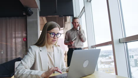 pleasant-looking woman works on her laptop at table. man with paper book in his hand talks on the phone at backdrop.