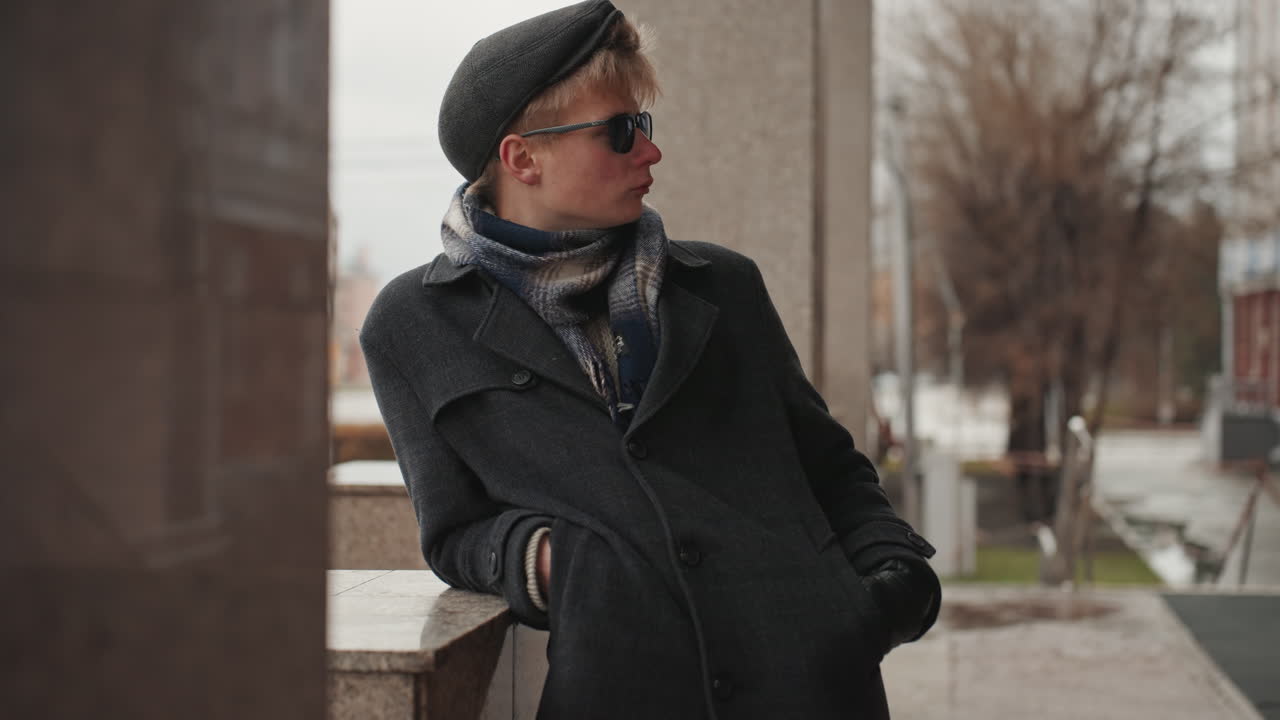 Angolo di città con un creatore bianco che assume una posa casual, appoggiato a un davanzale in pietra con occhiali da sole e sciarpa, atmosfera da servizio fotografico di moda urbana con pioggia sottile, sfondo con cartellone pubblicitario e pini, energia da influencer.