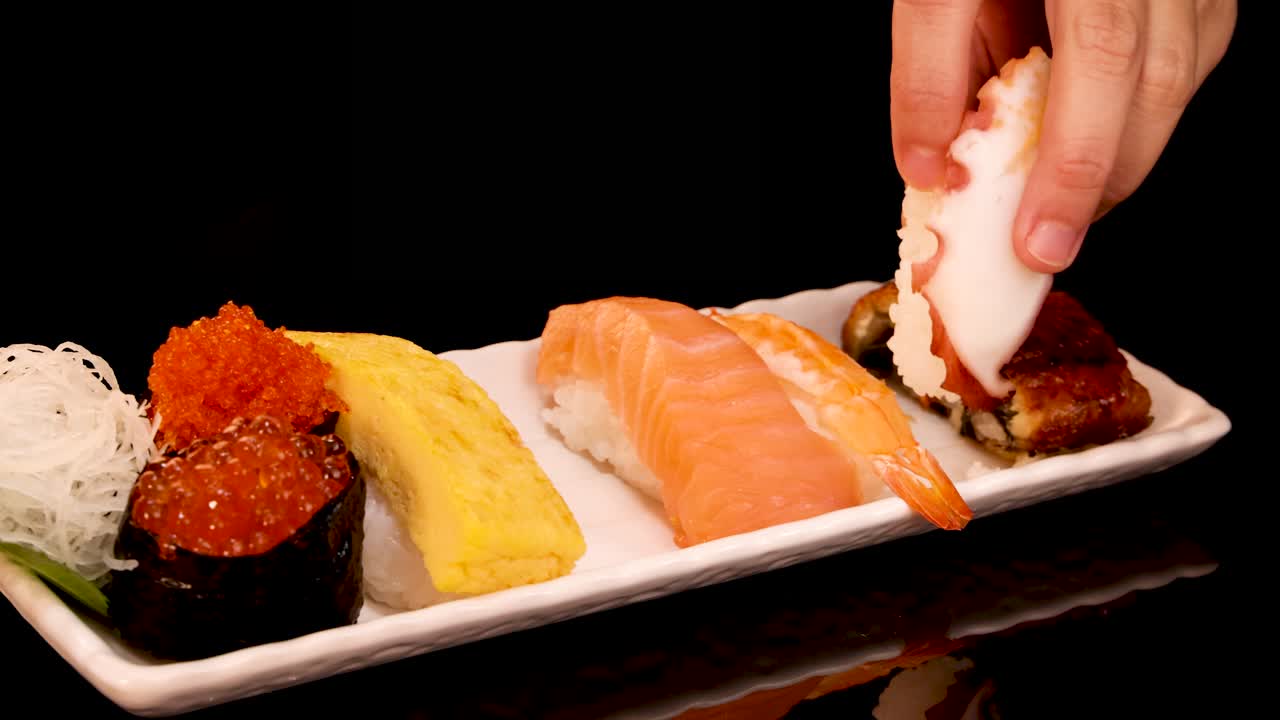 A hand picks various nigiri and gunkan sushi pieces from a neatly arranged plate under warm, studio lighting, with smooth camera movement and close-up framing