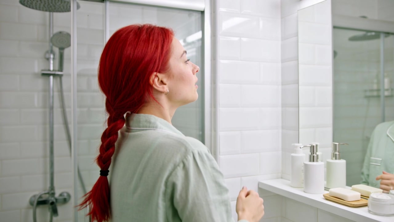 Touching woman with red braid in green pajama top checking jawline facing bathroom mirror and pumps