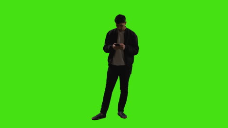 joven enviando o leyendo un mensaje de texto o desplazándose en línea en un teléfono móvil de pie contra una pantalla verde con iluminación de bajo nivel