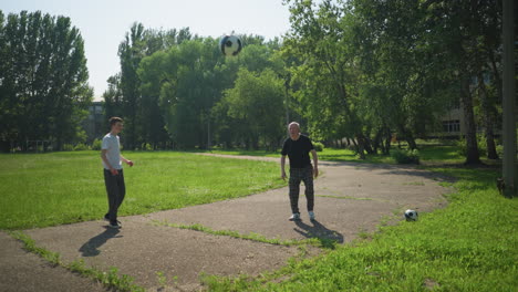 an elderly man skillfully heads a ball thrown to him while standing on a walkway, with his grandson observing nearby, with ball in the background and trees around