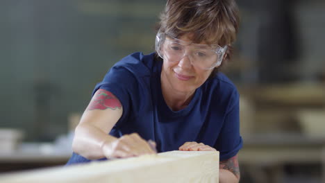 woman in safety goggles sanding timber with sandpaper and then blowing dust away