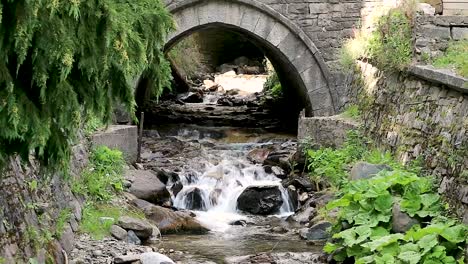 Historical-old-stone-bridge-over-streaming-water-in-forestry-area-near-Rila-Monastery,-zoom-in-shot