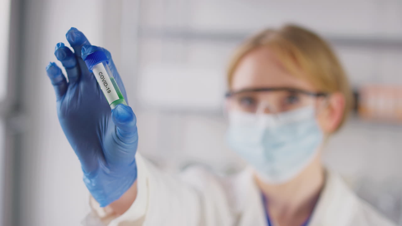 trabajadora de investigación de laboratorio con gafas de seguridad y máscara sosteniendo un tubo de ensayo etiquetado covid-19