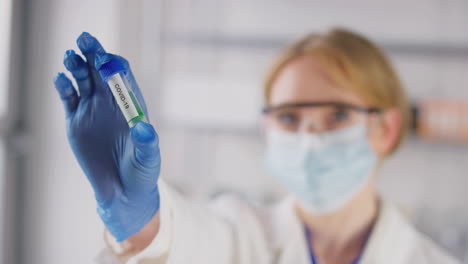 female lab research worker wearing safety glasses and mask holding test tube labelled covid 19