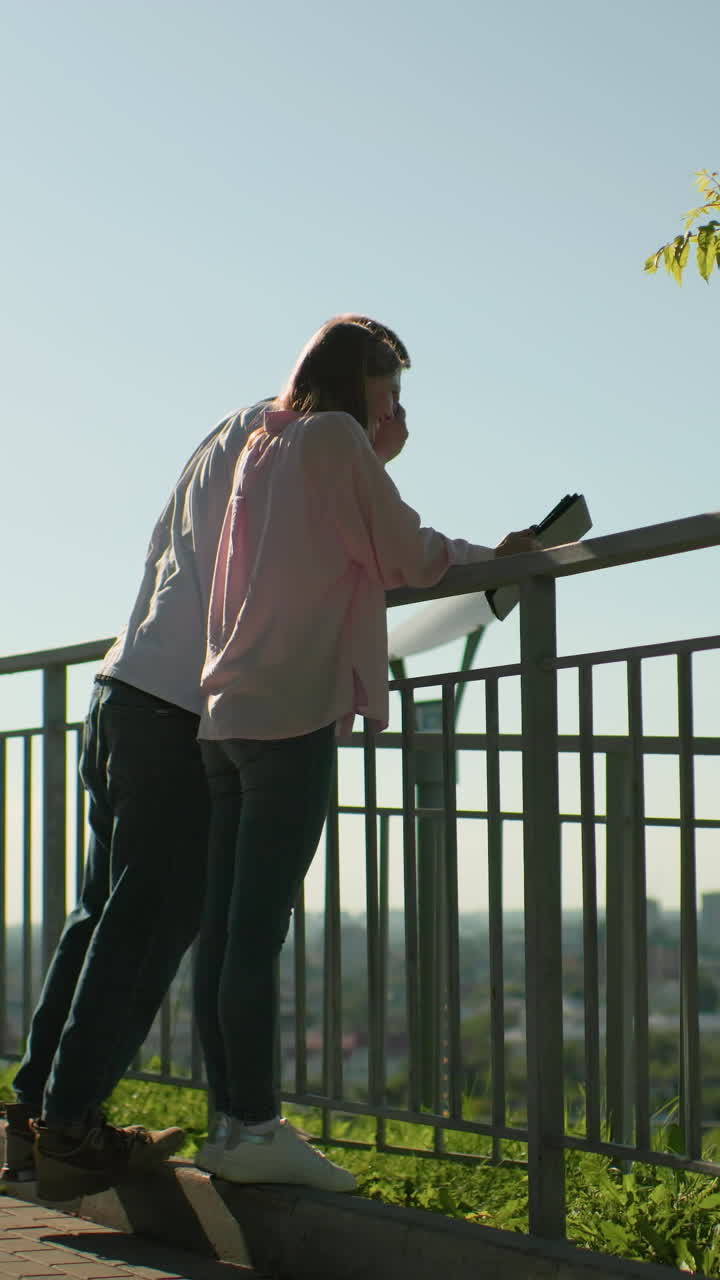 vista lateral de amigos apoyados en una barandilla de hierro mientras el niño se ríe cubriendo su cara mientras la dama muestra algo en la tableta, el fondo presenta un paisaje urbano vibrante, vegetación y atmósfera soleada