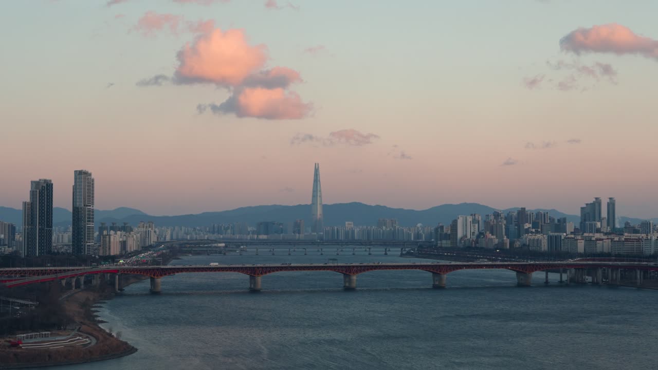 Vista aerea dello skyline di Seoul con la Lotte World Tower e il ponte Seongsu sul fiume Han al tramonto, soffici nuvole rosa e il traffico cittadino sottostante in una serata invernale