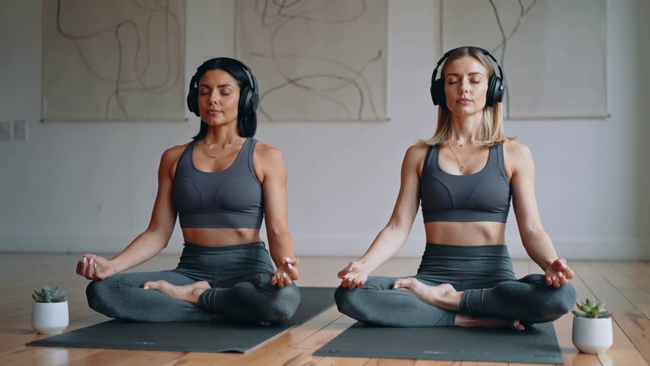 Duas mulheres meditando com foco na respiração para a calma em estúdio, com tapetes de ioga, fones de ouvido e roupas cinza combinando.