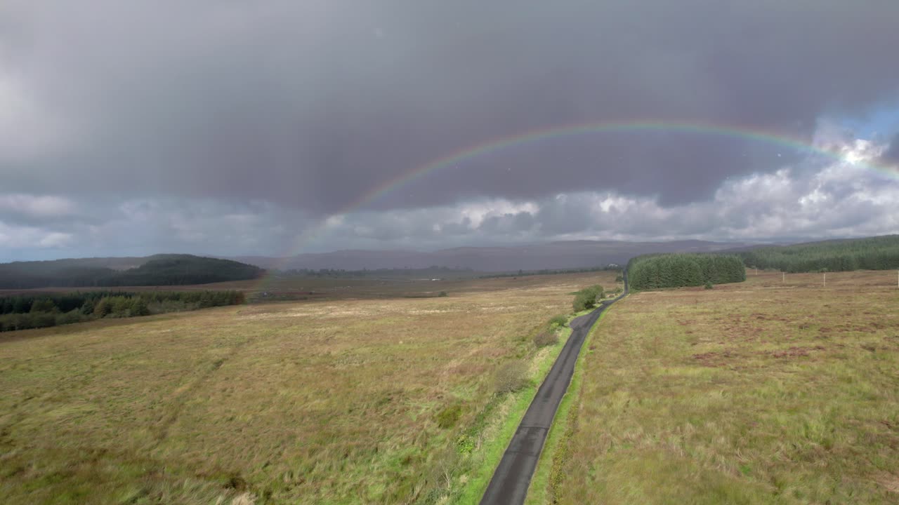 imágenes aéreas de drones bajo la lluvia que se elevan sobre un camino largo y recto mirando hacia un arco iris brillante con nubes grises oscuras en el fondo, con campos, árboles y páramos en escocia