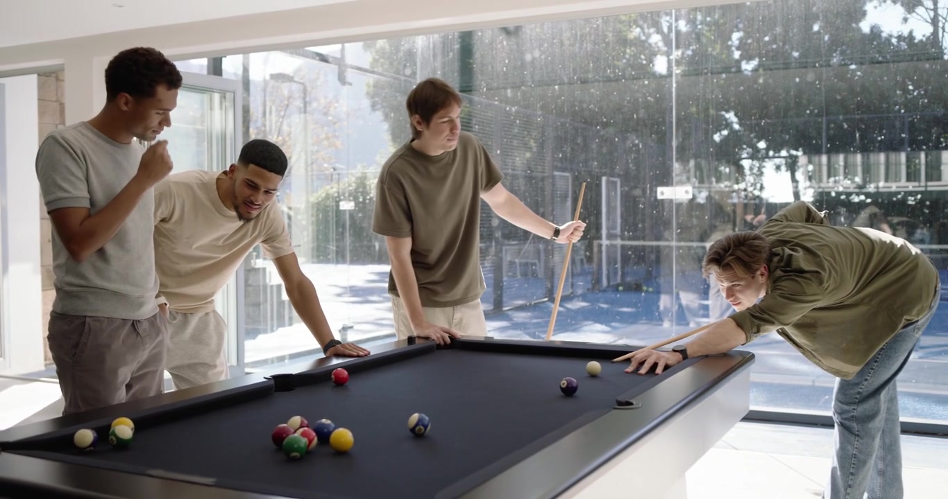 Diverse male friends in glass room playing pool, cue striking and scattering balls on black table