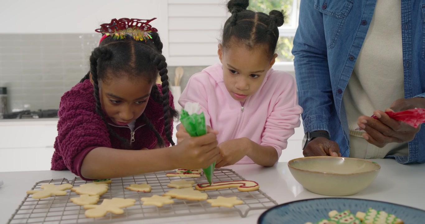 Uomo afroamericano che guida due bambine mentre decorano biscotti semplici con la glassa sul bancone per preparare dei dolcetti