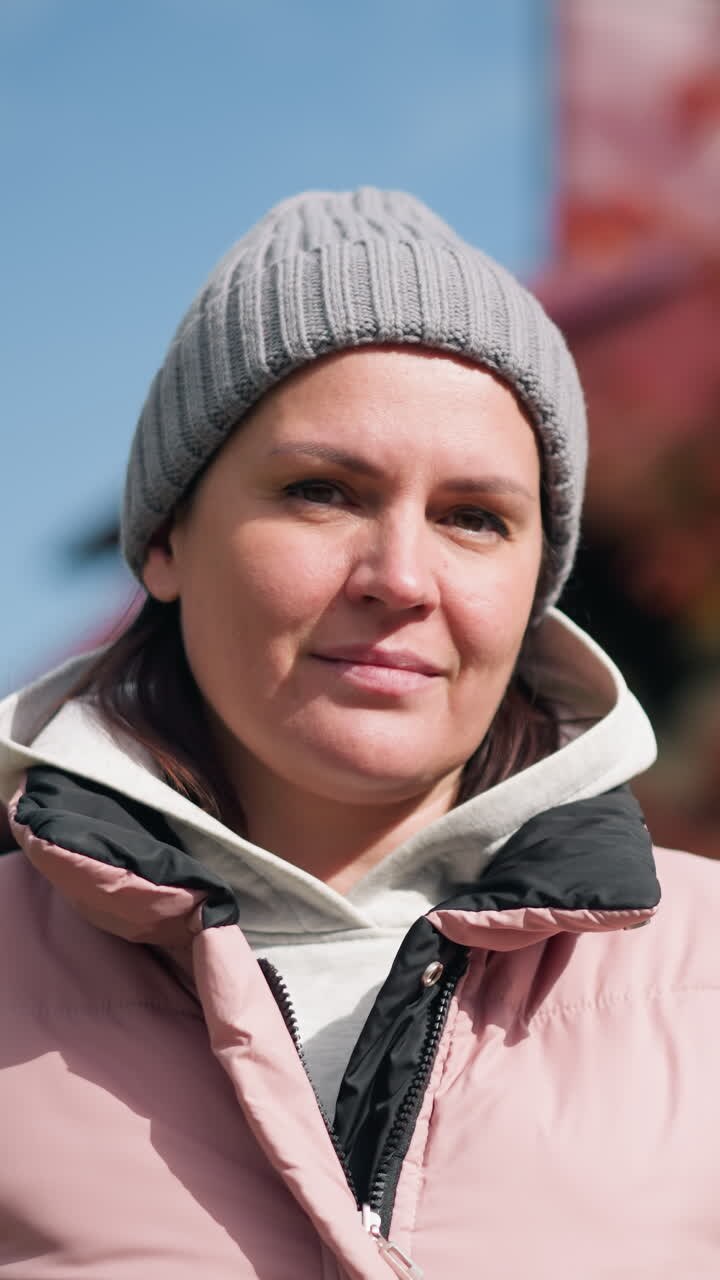 Giovane donna con giacca rosa e berretto grigio, con un'espressione pensierosa, che inclina la testa con un leggero battito di ciglia, all'aperto con alberi autunnali sfocati e una casa sullo sfondo sotto un cielo sereno.