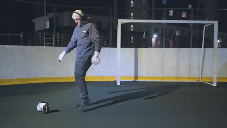 atleta practicando habilidades dinámicas de fútbol, equilibrando y pateando la pelota bajo la luz enfocada en la cancha al aire libre, entorno urbano con marcas de límite amarillas y esgrima en el fondo