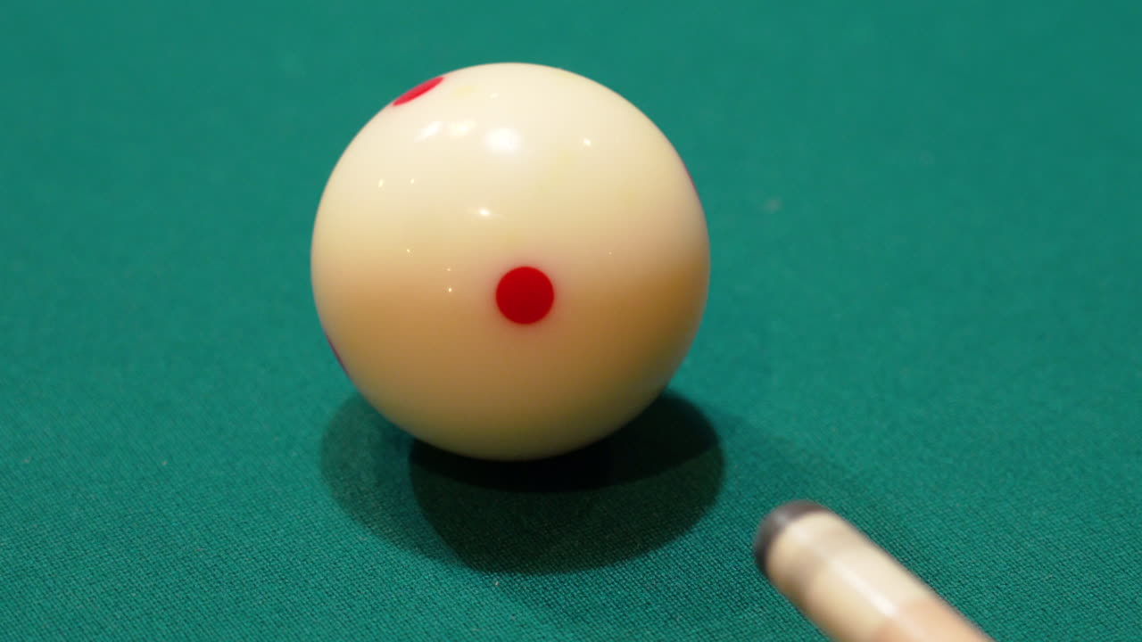 primer plano de billar de una bola blanca con manchas rojas disparadas en una mesa de billar con una punta de palo de madera que se clava en fieltro verde después de varios golpes de práctica, sin rostros