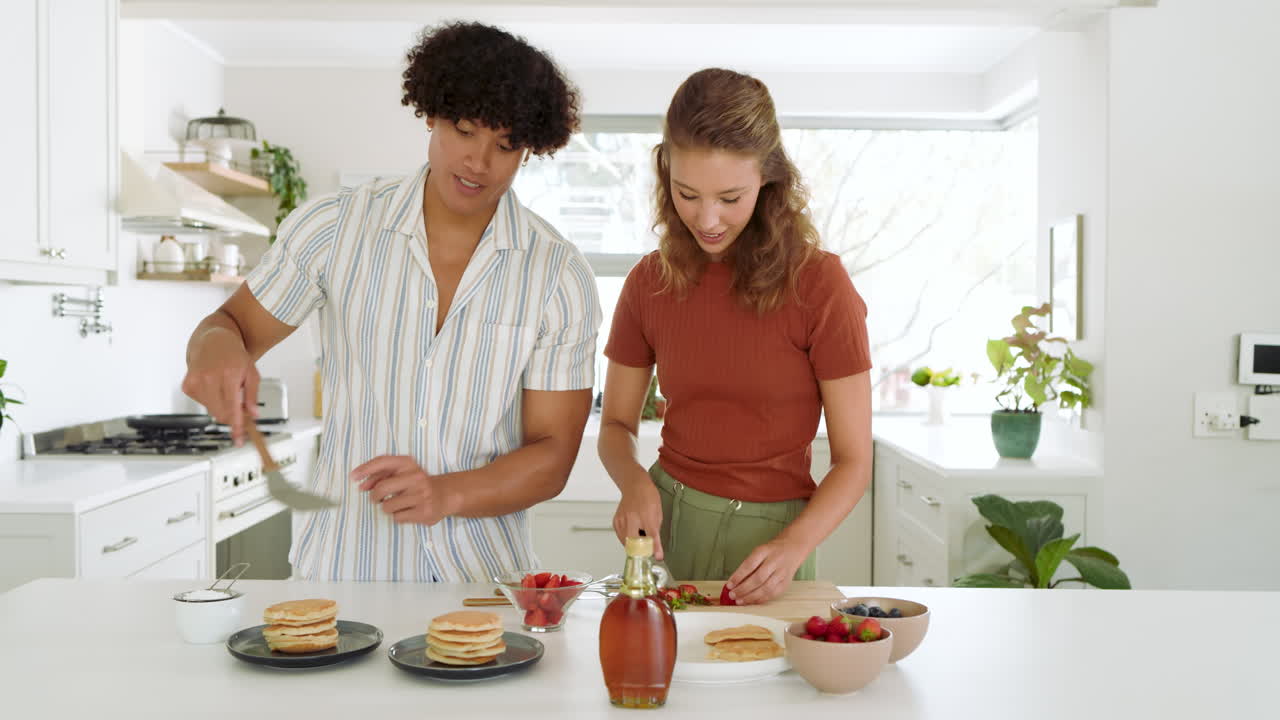 Preparing breakfast, diverse couple slicing strawberries and stacking pancakes in kitchen, at home
