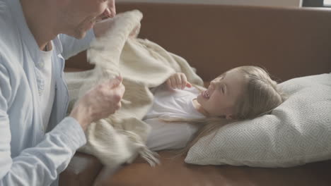 little daughter lying in the sofa, her funny dad covers her with a quilt