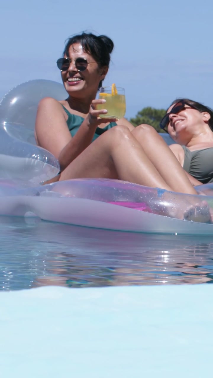 Vertical video: Sun warming women in swimsuits on float in private pool, raising glass