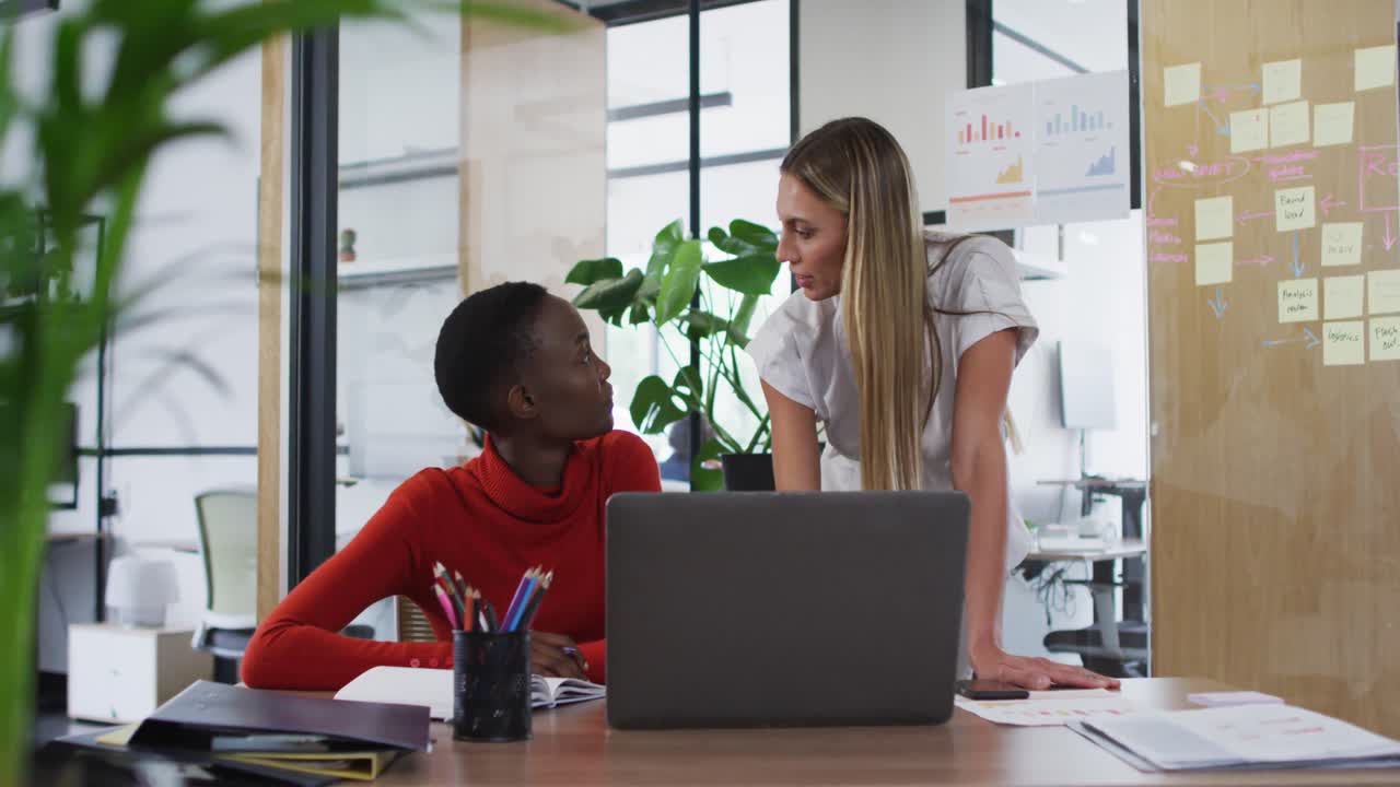 Two diverse female office colleagues with laptop discussing together at office