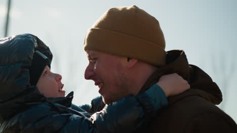 a close up of father carrying his son, both of them are conversing with each other happily as the father moved him playfully, they both are wearing jacket and a beanie