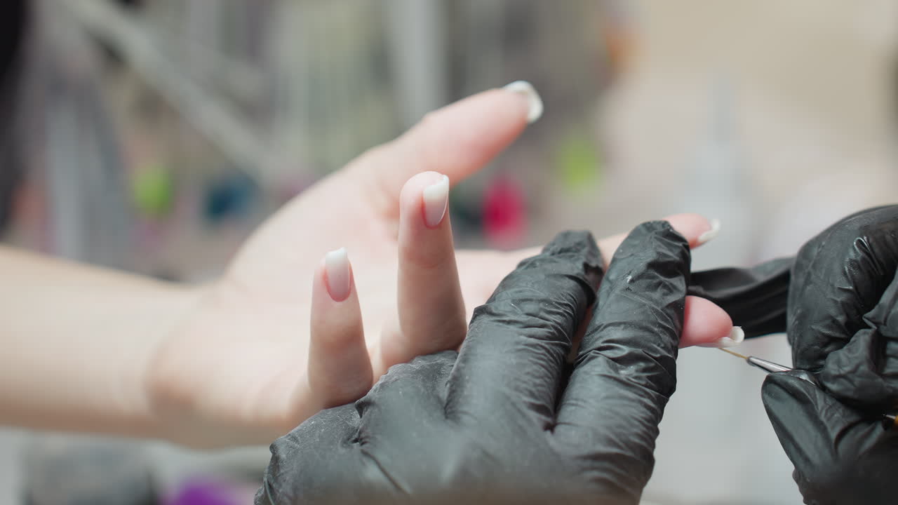 Nail technician in black gloves carefully applies white acrylic on client fingernail using precision brush, highlighting attention to detail and clean nail care environment with blurred background