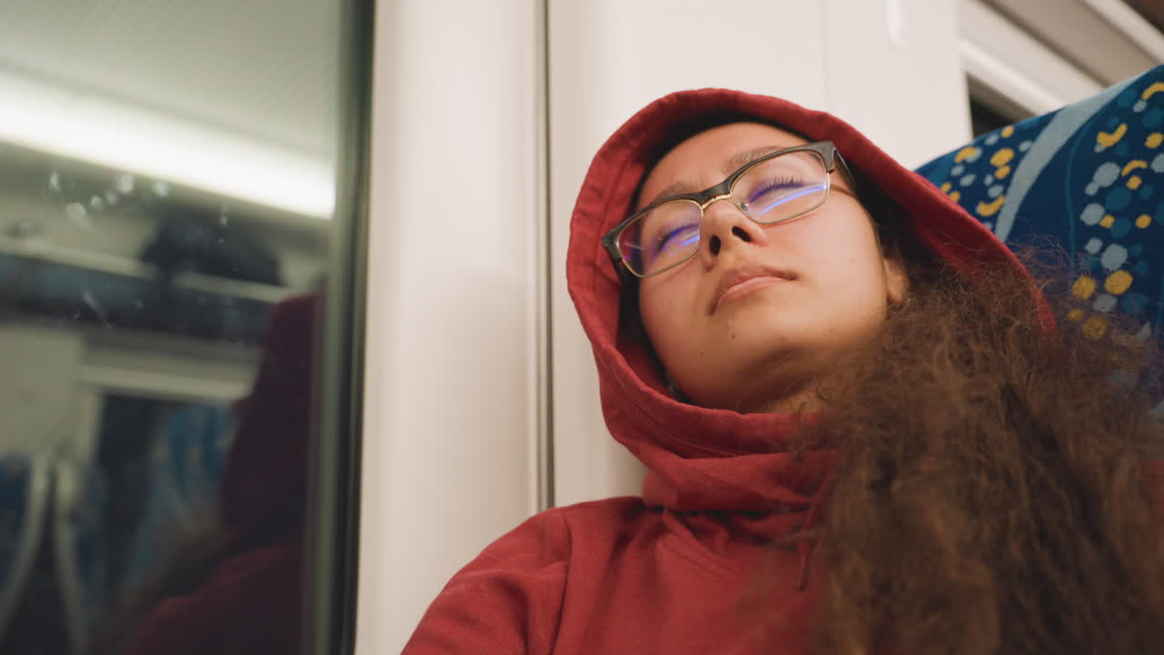 Giovane artista che sonnecchia accanto al finestrino del treno, in transito notturno con riflessi al neon, felpa rossa che incornicia il viso, occhiali che catturano le luci della città, testa inclinata verso il vetro, bokeh morbido, atmosfera introspettiva.