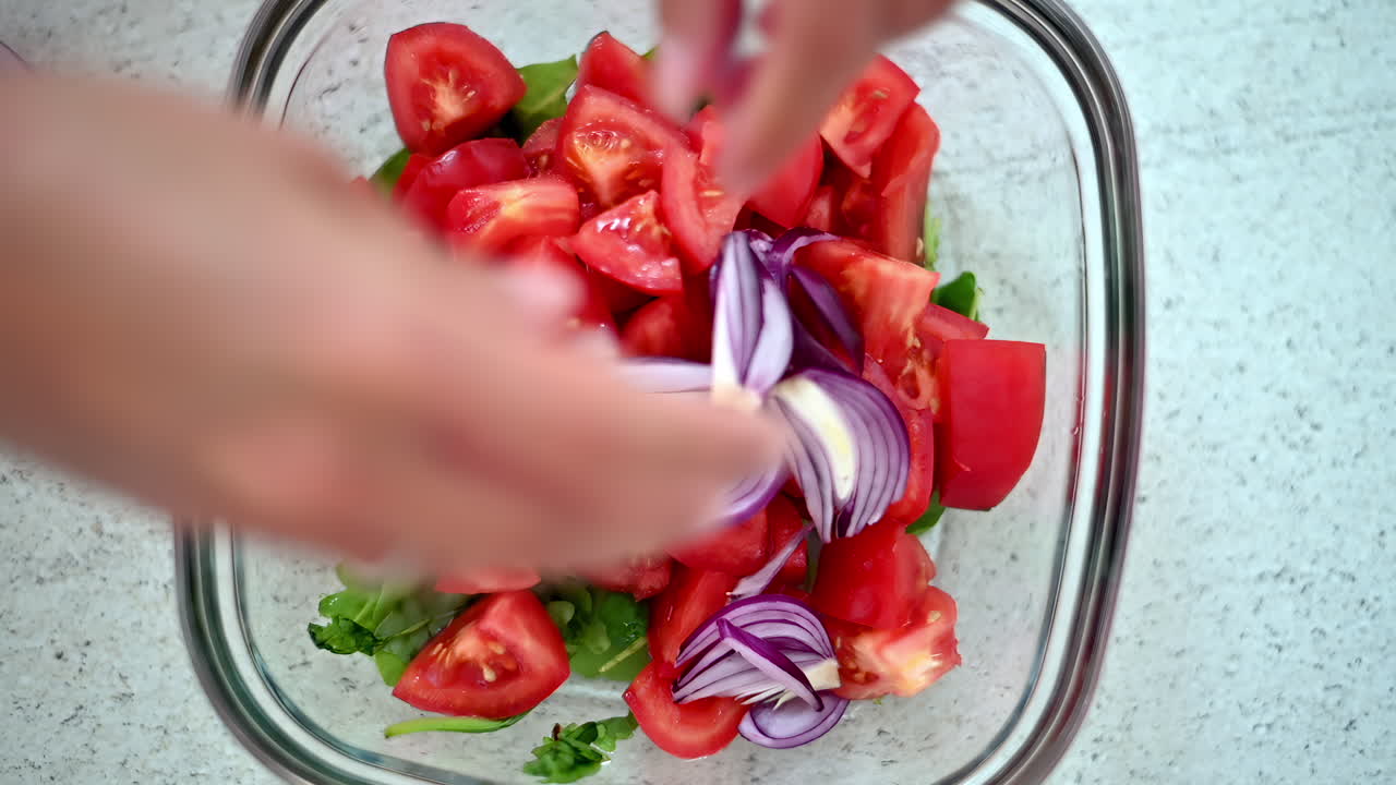 Hands prepare fresh tomatoes and purple onions for a colorful salad. The ingredients are carefully cut and mixed in a clear bowl, showcasing healthy cooking