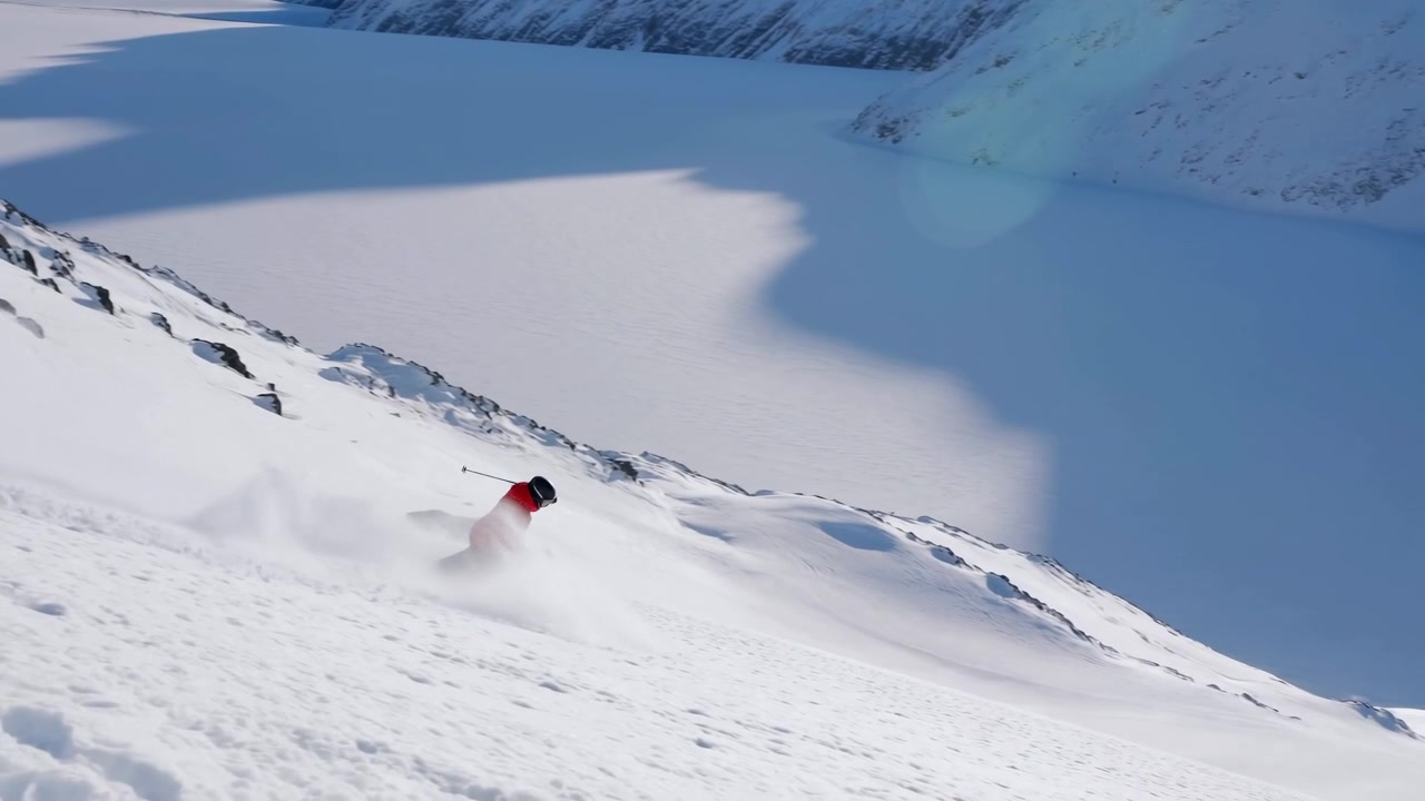 Skier in red jacket and black helmet carves powerful moving turns down steep powder slope, sending spray across granular snow with glacier valley, rocky ridge, bright sunlight, long shadows