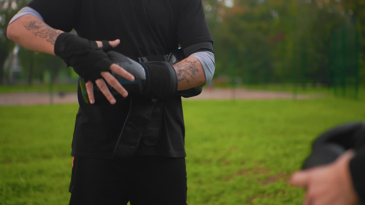 Nahaufnahme von Händen, die beim Parktraining Boxhandschuhe anpassen, Riemen befestigen, Klebeband überprüfen, tätowierte Unterarme, methodische Routine, Trainersilhouette im Hintergrund, Fokus auf Griff und Technik